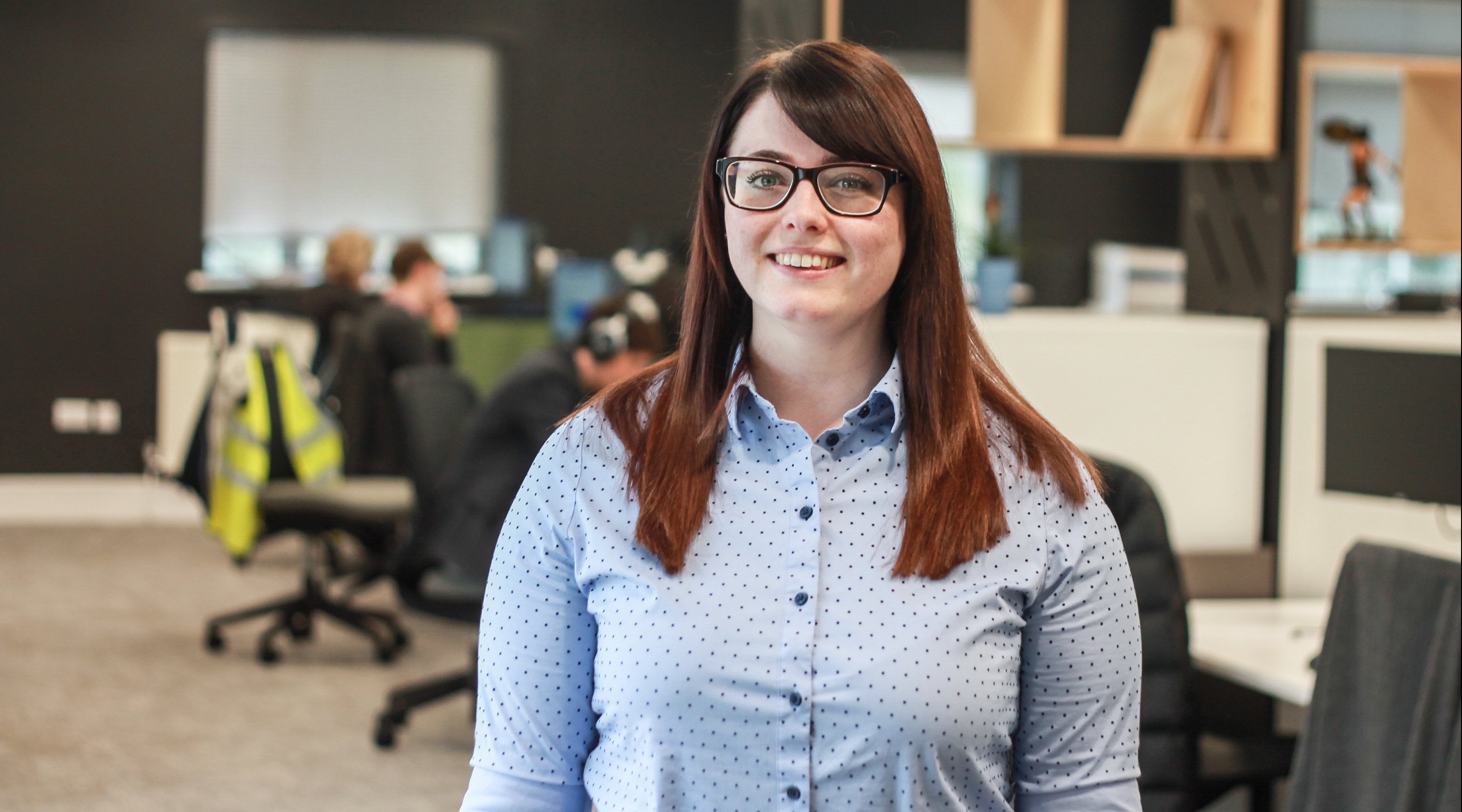 Brown haired woman with glasses and wearing a pale blue button up shirt standing in an office